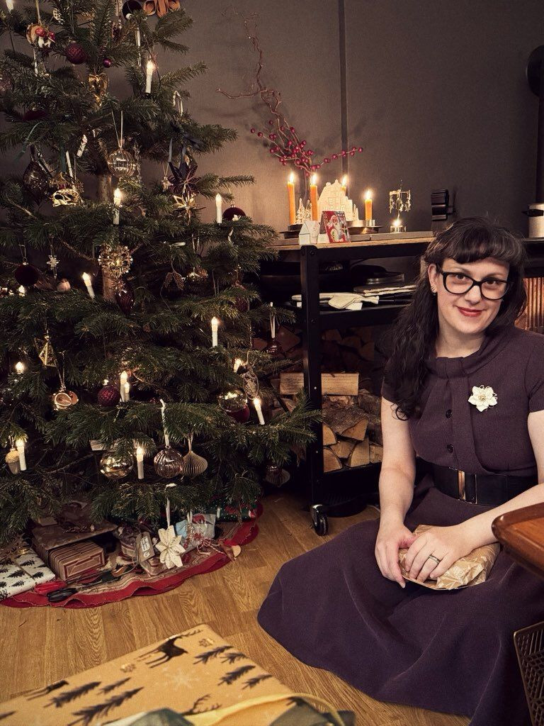image of a woman (me) with dark hair and glasses wearing mauve-burgundy dress, sitting next to an evergreen tree decorated with lights and Christmas ornaments